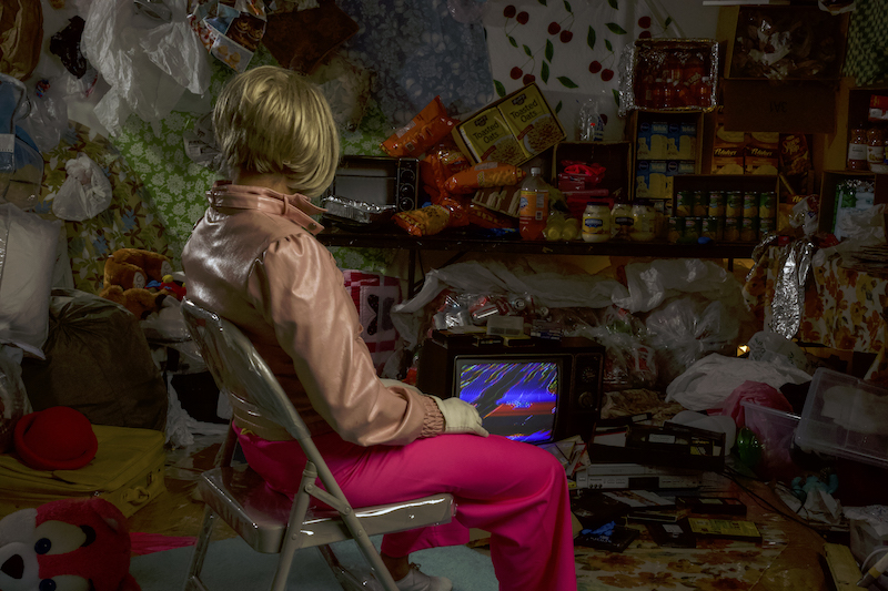 A performer in a blonde-bobbed wig stares at a grainy old TV that's in midst of a pile of junk.